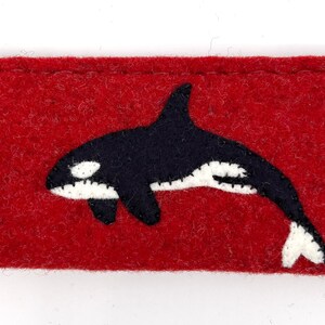 Op de afbeelding: Een rechthoekig etui van rode vilt met een applicatie van een zwart-witte orka in het midden. Het etui heeft zichtbare stiksels langs de randen en een zacht, getextureerd oppervlak, wat dient als een platte beschermhoes voor kleine items.