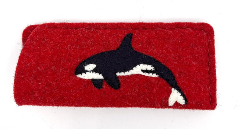 Op de afbeelding: Een rechthoekig etui van rode vilt met een applicatie van een zwart-witte orka in het midden. Het etui heeft zichtbare stiksels langs de randen en een zacht, getextureerd oppervlak, wat dient als een platte beschermhoes voor kleine items.