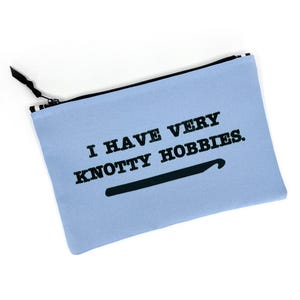 May include: A light blue rectangular zippered pouch featuring the black printed text "I HAVE VERY KNOTTY HOBBIES." accompanied by a black graphic of a crochet hook. The pouch has a black zipper closure with a black and white striped pull tab.