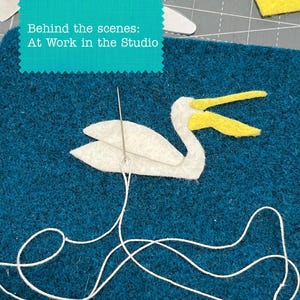 May include: A white felt pelican applique with a yellow beak is positioned on a dark teal felt fabric. A silver sewing needle is inserted into the pelican, with a long white thread trailing down across the textured blue surface below.