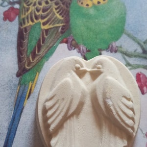 May include: A heart-shaped, off-white decorative item featuring two birds facing each other, with their wings spread. The background includes a watercolor painting of two parakeets perched on a branch with pink flowers.