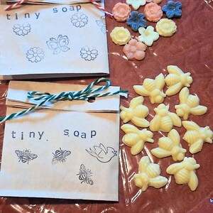 May include: Two small paper packages labeled "tiny soap" with floral and insect illustrations, tied with twine. Scattered around are small, flower-shaped soaps in various pastel colors and yellow bee-shaped soaps, all on a reddish-brown surface.