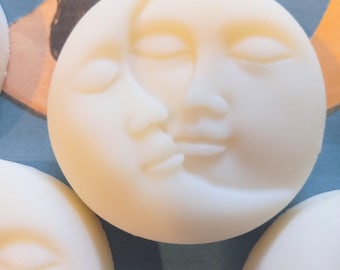 May include: Close-up of several off-white, circular soaps. Each soap features a detailed relief of a crescent moon kissing a face. The soaps are arranged on a blue and green background. The soaps have a smooth, matte finish.