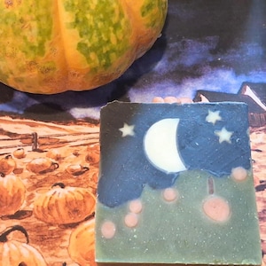 Puede incluir: Una pastilla de jabón cuadrada que presenta un cielo nocturno azul oscuro con una luna creciente blanca y pequeñas estrellas blancas sobre un paisaje verde con acentos circulares de color rosa claro. El jabón tiene una superficie plana.