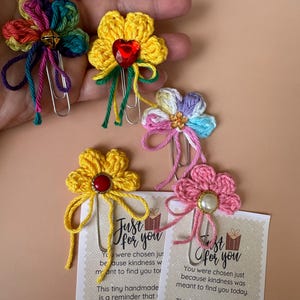 May include: Crocheted flower paper clips in various colors, including yellow, pink, and rainbow hues. Each clip features a decorative center, such as a gem or bead, and a dangling yarn accent. The clips are paired with cards that read "Just for you."