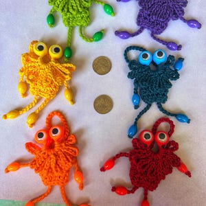 May include: A collection of six handmade crochet monster figures in various colors: green, purple, yellow, teal, orange, and red. Each monster has large googly eyes and beaded limbs. The figures are displayed on a white surface.