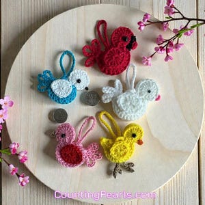 May include: A collection of five colorful, crocheted bird ornaments. The birds are in shades of red, blue, white, pink, and yellow, with black and orange accents. Each bird has a loop for hanging. The ornaments are displayed on a wooden surface.