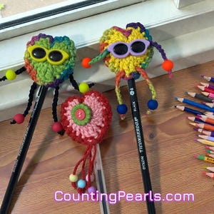 May include: Handmade crochet heart pencil toppers with sunglasses and beaded accents. The toppers are attached to black pencils. The pencils are decorated with colorful yarn and beads. The image includes the text "CountingPearls.com".