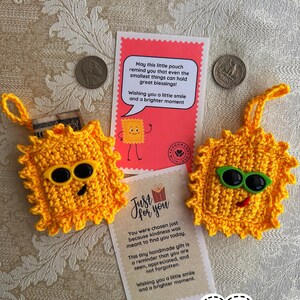 May include: Two handmade yellow crochet sun-shaped pouches with black sunglasses and smiling faces. The pouches are next to cards with text about kindness and a brighter moment. Two coins are also visible.