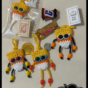 Puede incluir: Una colección de figuras de ganchillo hechas a mano con cuerpos amarillos, naranjas y blancos, extremidades de cuentas negras y gafas de sol. La imagen incluye una barra de chocolate Snickers y Twix, un pequeño libro y una etiqueta de regalo gratis. También es visible el canal de YouTube "CountingArtsNCrafts".