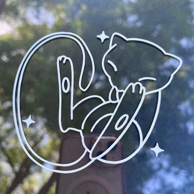 May include: White outline of a cartoon cat-like creature in a curled pose, with three small star shapes around it. The design is on a dark surface, possibly a window or sticker.