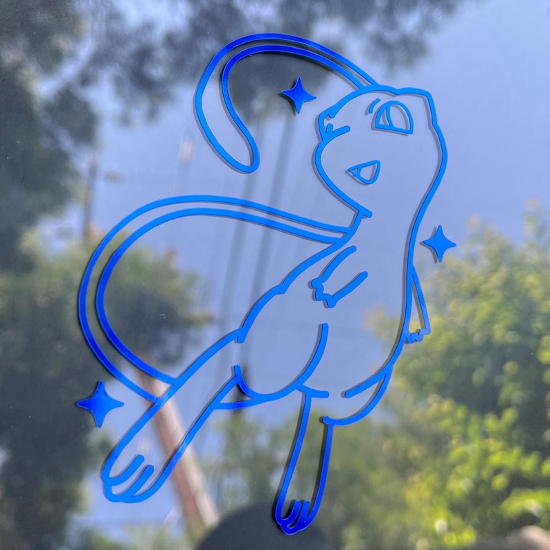 May include: A blue and white decal of the Pok&eacute;mon Mew, with a stylized design. The decal features a smiling Mew with a long tail and three star accents. The image is set against a blurred background of a window and trees.
