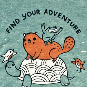 May include: A teal blue t-shirt with a graphic of a turtle, a beaver, a frog, and two birds. The text "Find Your Adventure" is written in a black, curved font around the graphic. The text "Susie Ghanretmon/ BoyGirlParty for Artisans" is printed in black at the bottom of the graphic.