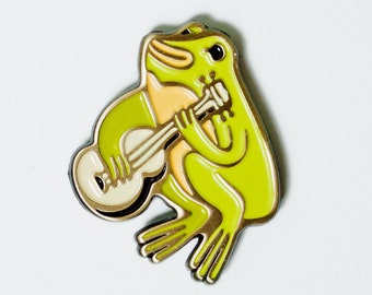 Puede incluir: Un pin de esmalte dorado con una rana verde tocando el ukelele. El pin dice "ukulele frog pin" y "susie ghahremani / boygirlparty®".