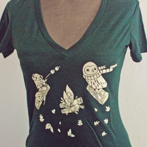 May include: A dark green t-shirt with a white graphic of owls around a campfire. The text "susie ghahremani / boygirlparty" is printed on the shirt.