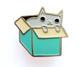 May include: A grey cat enamel pin with gold accents. The cat is peeking out of a teal box with a yellow top. The pin says "grey cat pin" at the top and "susie ghahremani / boygirlparty®" at the bottom.