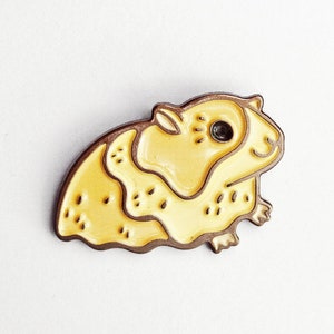 May include: A yellow enamel pin of a guinea pig. The pin has a brown outline and a black eye. The text "guinea pig pin" is above the pin.