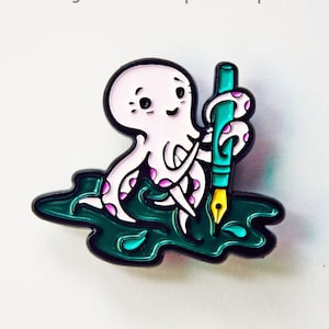May include: A pink octopus with purple spots holding a teal pen with a gold nib. The octopus is sitting in a pool of green ink with a few ink drops around it. The text "inky octopus pin" is above the octopus and the text "susie ghahremani / boygirlparty®" is below the octopus.