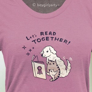 May include: A pink t-shirt featuring a white dog and cat reading a book together. The text on the shirt reads "Let's Read Together!"