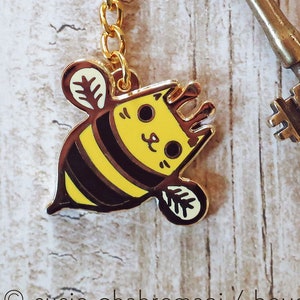May include: A gold keychain with a cute cartoon cat bee charm. The charm is yellow and black with a cat face and bee wings. The keychain also includes a vintage-style key.