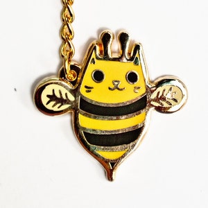 May include: Gold keychain featuring a cartoon cat bee design. The bee has black stripes, yellow wings, and a cute cat face. The keychain is attached to a gold ring and chain.