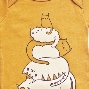 May include: A mustard yellow baby onesie with a graphic of a stack of four white cats with black eyes and brown outlines. The text "susie ghahremani / boygirlparty&reg;" is at the bottom of the onesie.