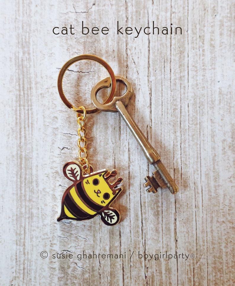 May include: A gold keychain with a cute cartoon cat bee charm. The charm is yellow and black with a cat face and bee wings. The keychain also includes a vintage-style key.