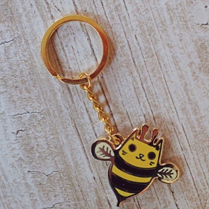 May include: A gold keychain with a bee charm. The bee is yellow and black with a cat face. The text "susie ghahremani / boygirlparty&reg;" is visible in the top left corner of the image.