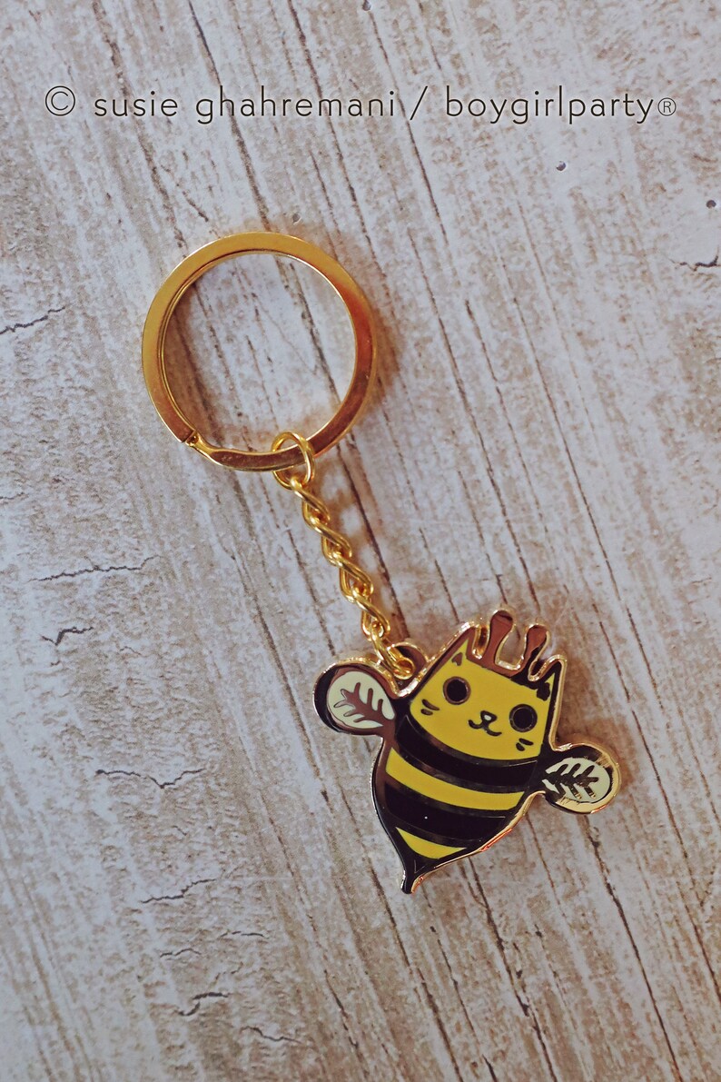 May include: A gold keychain with a bee charm. The bee is yellow and black with a cat face. The text "susie ghahremani / boygirlparty&reg;" is visible in the top left corner of the image.