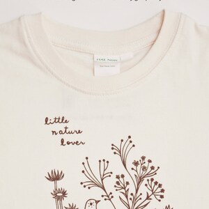 May include: A cream colored t-shirt with a brown graphic of a bird and flowers. The text "little nature lover" is printed above the graphic.
