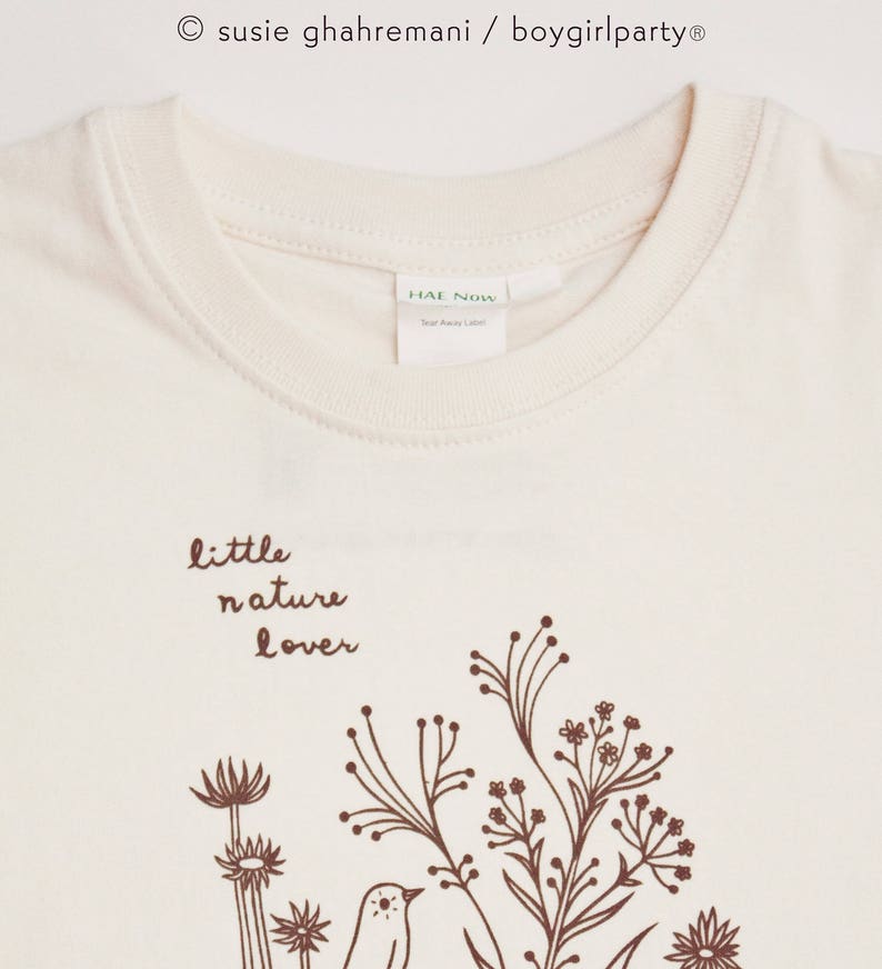 May include: A cream colored t-shirt with a brown graphic of a bird and flowers. The text "little nature lover" is printed above the graphic.