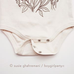 Puede incluir: Un body blanco con un dibujo floral en l&iacute;nea marr&oacute;n. El body tiene un cierre de presi&oacute;n en la entrepierna y una firma "susie ghahremani / boygirlparty&reg;" en la parte inferior.