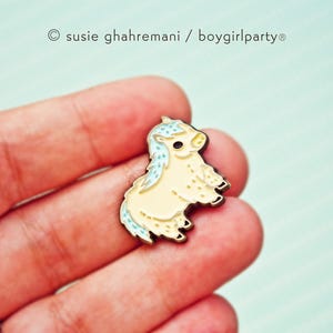 Op de afbeelding: Een kleine, gele emaille pin van een cartoon pony met een blauwe manen en staart. De pony staat op alle vier poten en heeft een schattige uitdrukking op zijn gezicht.