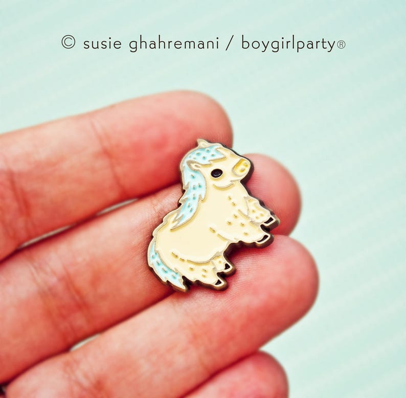 Op de afbeelding: Een kleine, gele emaille pin van een cartoon pony met een blauwe manen en staart. De pony staat op alle vier poten en heeft een schattige uitdrukking op zijn gezicht.