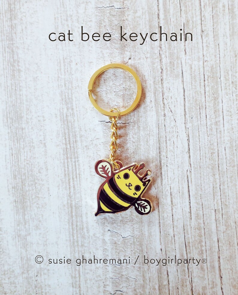 May include: Gold keychain featuring a cartoon cat bee with black and yellow stripes. The keychain has the text "susie ghahremani / boygirlparty&reg;" at the bottom.