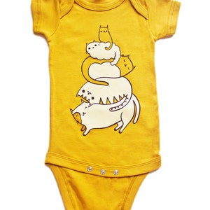 May include: A mustard yellow baby onesie with a graphic of a stack of five white cats with black eyes and brown outlines. The text "susie ghahremani / boygirlparty&reg;" is at the bottom of the onesie.