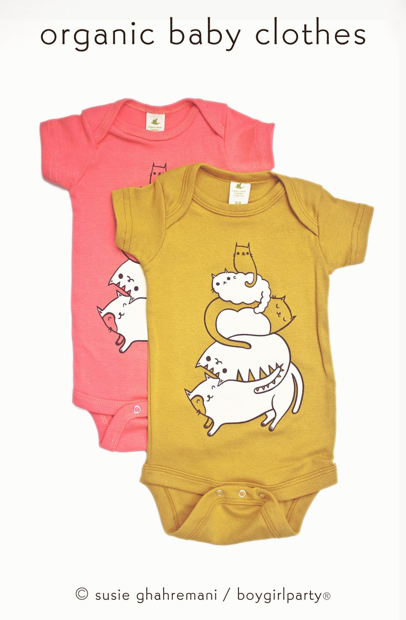 May include: Two organic cotton baby onesies. One is pink with a cat graphic. The other is mustard yellow with a stack of cats graphic. The text "susie ghahremani / boygirlparty&reg;" is at the bottom of the image.