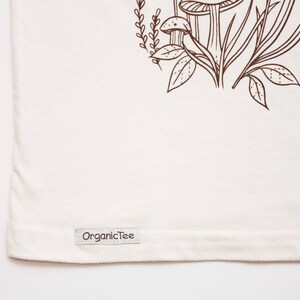 May include: A white t-shirt with a brown line drawing of mushrooms and plants. The shirt has a brown label that says "OrganicTee" on the bottom.