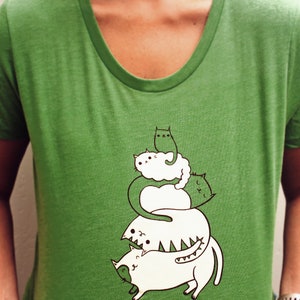 May include: Green t-shirt with a graphic of four white cats stacked on top of each other. The cats are drawn in a cartoon style and have different expressions. The text "susie ghahremani / boygirlparty&reg;" is printed at the top of the shirt.