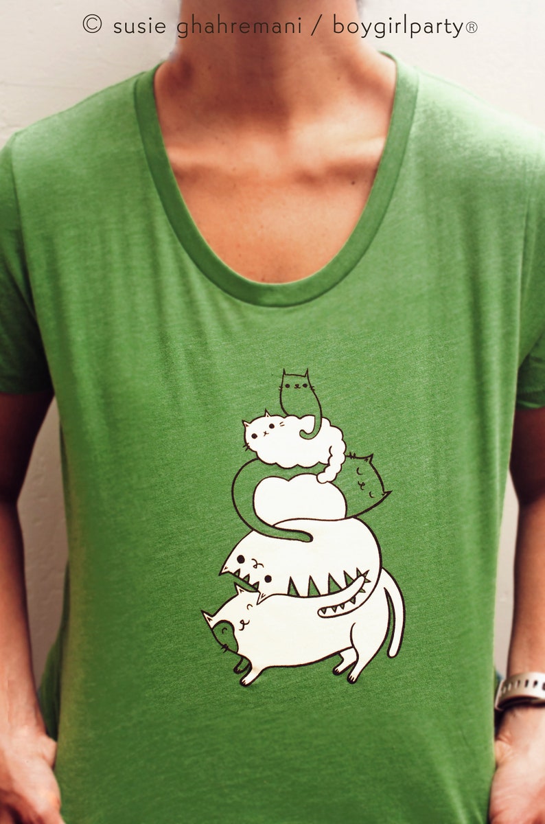 May include: Green t-shirt with a graphic of four white cats stacked on top of each other. The cats are drawn in a cartoon style and have different expressions. The text "susie ghahremani / boygirlparty&reg;" is printed at the top of the shirt.