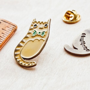 May include: A gold-colored enamel pin of a cat wearing a green bow tie. The cat is sitting up with its tail curled around its body. The pin is on a wooden surface. The back of the pin is silver and has the words "BoyGirlParts" engraved on it.