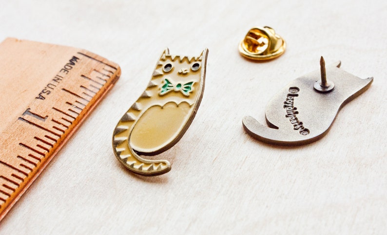 May include: A gold-colored enamel pin of a cat wearing a green bow tie. The cat is sitting up with its tail curled around its body. The pin is on a wooden surface. The back of the pin is silver and has the words "BoyGirlParts" engraved on it.
