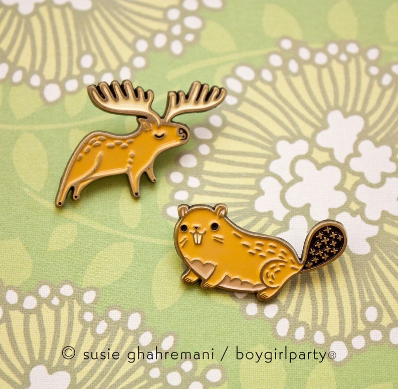 May include: Two gold enamel pins featuring a yellow moose and a yellow beaver. The pins are on a green and white floral background.