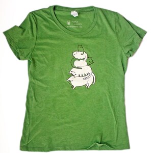 May include: Green eco-friendly t-shirt with a graphic of a stack of five white cats with green accents. The text "susie ghahremani / boygirlparty&reg;" is at the bottom of the shirt.