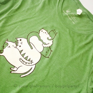 May include: Green t-shirt with a white graphic of a stack of cats. The graphic features five cats, each with a different expression. The text "susie ghahremani / boygirlparty" is printed at the bottom of the graphic.