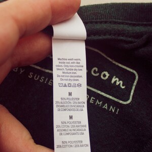 May include: A white care label with washing instructions for a green shirt. The label includes the text "Machine wash warm, inside out, with like colors. Only non-chlorine bleach. Tumble dry low. Medium iron. Do not iron decoration. Do not dry clean." The label also includes the size "M" and the fabric composition: "50% Polyester, 25% Cotton, 25% Rayon". The label states that the shirt was assembled in Nicaragua from components made in the USA.