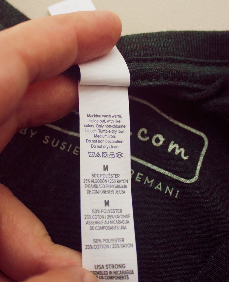May include: A white care label with washing instructions for a green shirt. The label includes the text "Machine wash warm, inside out, with like colors. Only non-chlorine bleach. Tumble dry low. Medium iron. Do not iron decoration. Do not dry clean." The label also includes the size "M" and the fabric composition: "50% Polyester, 25% Cotton, 25% Rayon". The label states that the shirt was assembled in Nicaragua from components made in the USA.