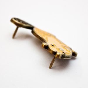 May include: A gold-toned enamel pin featuring a cartoon-style cat with a yellow body and black eyes. The pin has a back pin for attaching to clothing or a bag.