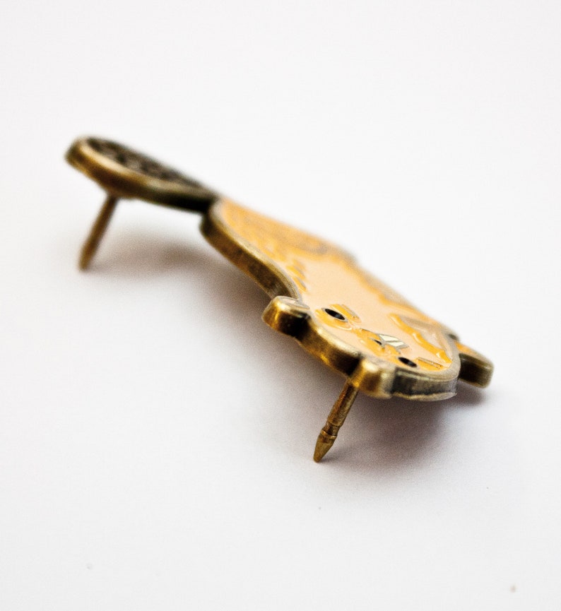 May include: A gold-toned enamel pin featuring a cartoon-style cat with a yellow body and black eyes. The pin has a back pin for attaching to clothing or a bag.
