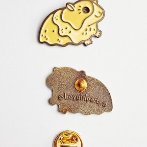 May include: A yellow enamel pin of a guinea pig with a brown outline. The back of the pin is brown with the text "&copy;boygirlparty" in white. The pin comes with a gold metal pin back.